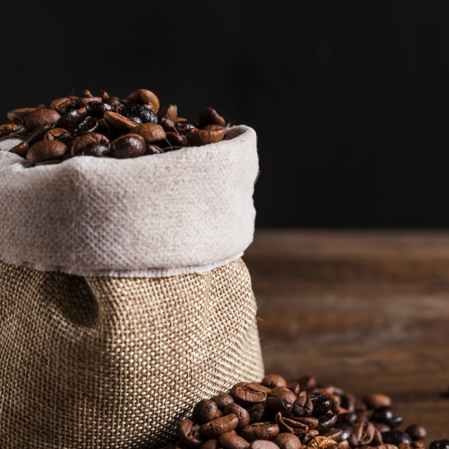 Burlap sack filled with roasted coffee beans, with additional beans spilled onto a wooden surface against a dark background.
