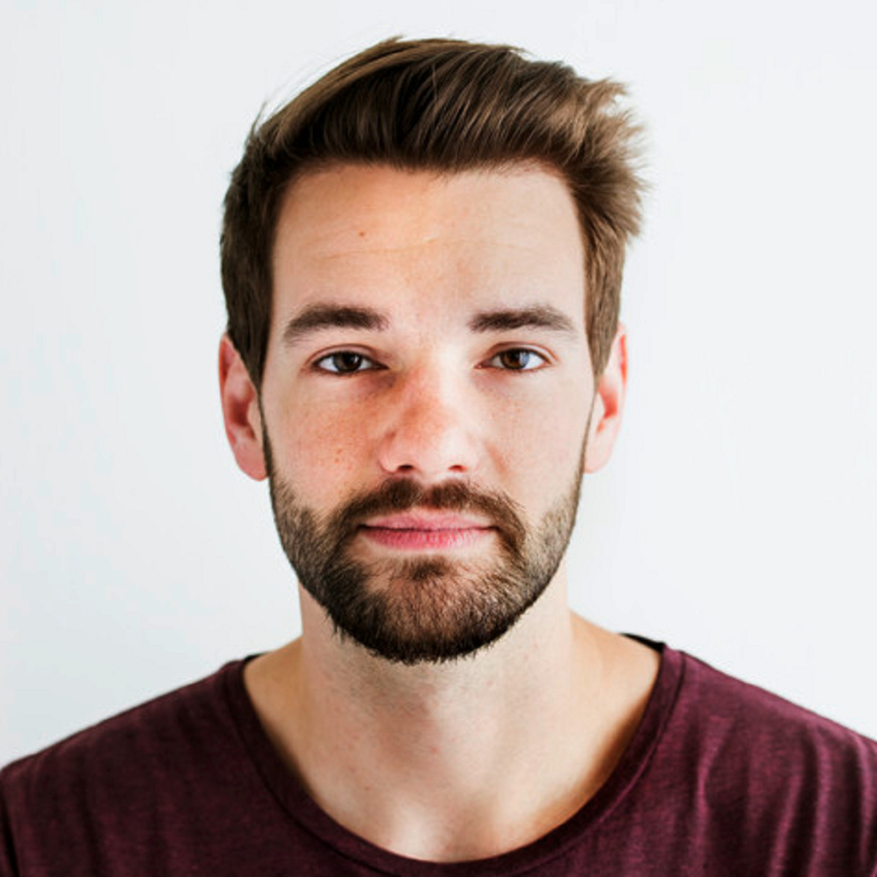 Man with short brown hair and a trimmed beard wearing a maroon shirt, photographed against a white background.