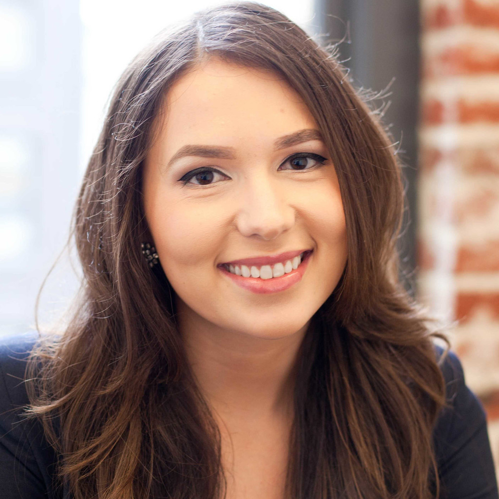 Smiling woman with long brown hair wearing a dark blazer, photographed indoors with a softly blurred background.