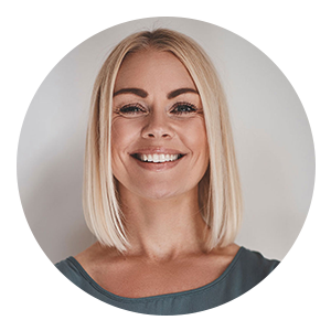 Circular portrait of a smiling woman with straight blonde hair cut to shoulder length, wearing a sleeveless top, facing the camera against a light neutral background.