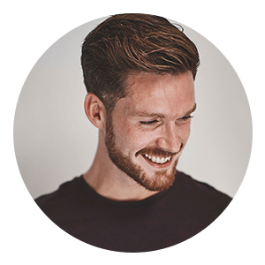 Circular portrait of a smiling man with short brown hair and a trimmed beard, looking slightly downward, wearing a dark shirt against a light neutral background.