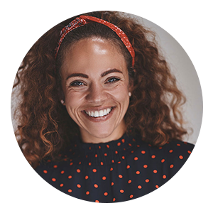 Circular portrait of a smiling woman with curly hair, wearing a red headband and a dark top with small red polka dots, photographed against a neutral background.
